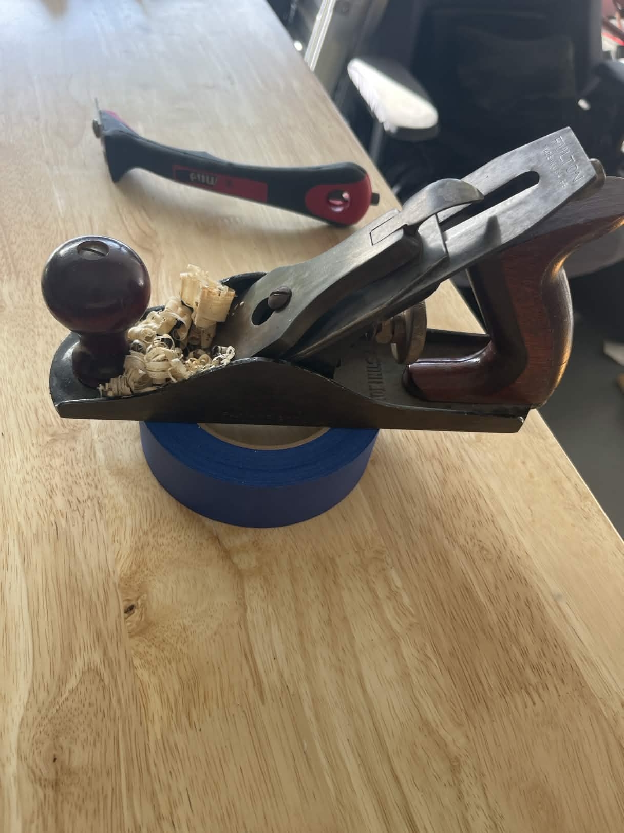 A Fulton No. 3097 hand plane. It has dark red wood for the fore and aft handles, the steel shoe is polished, and it sits on top of a roll of blue painters tape. All of this is on a wooden workbench.